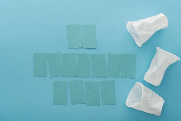 Vue du dessus des gobelets en plastique blanc et des notes collantes vides avec espace de copie sur fond bleu — Photo de stock