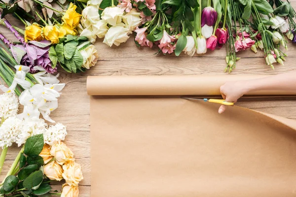 Partial view of florist cutting craft paper with scissors near flowers on wooden table — Stock Photo