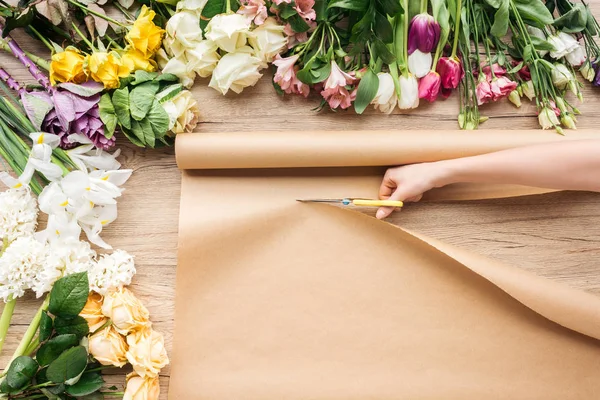 Partial view of florist cutting craft paper with scissors near flowers on wooden surface — Stock Photo