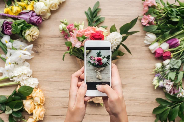 Vista cortada do florista segurando smartphone e tirando foto de buquê de flores na superfície de madeira — Fotografia de Stock