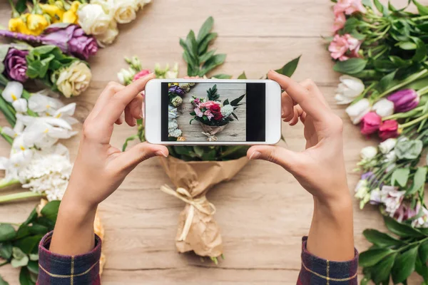 Vista recortada del florista sosteniendo el teléfono inteligente y tomando una foto del ramo de flores en la superficie de madera - foto de stock