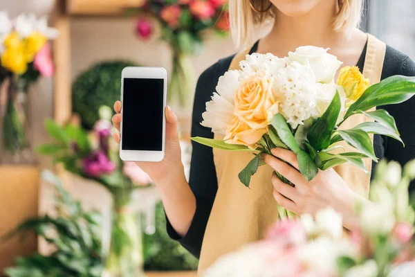 Vista recortada de floristería sosteniendo ramo de flores y mostrando el teléfono inteligente con pantalla en blanco - foto de stock