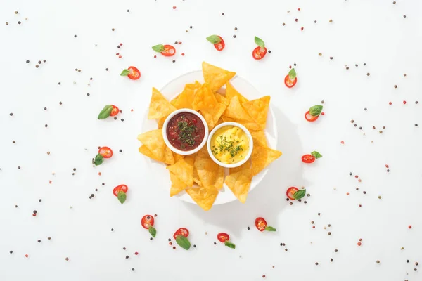 Vista superior de sabrosos nachos y salsas en plato, especias y chiles en rodajas con hojas de albahaca - foto de stock