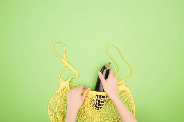 Cropped view of woman putting eggplant in yellow string bag on light green background — Stock Photo