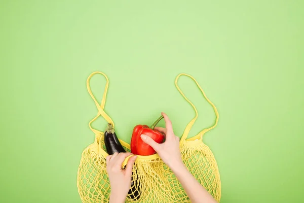 Cropped view of female hands, red bell pepper and eggplant in yellow string bag on light green background — Stock Photo