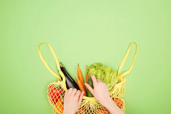 Vue partielle de la femme prenant du concombre frais dans un sac à ficelle jaune avec des légumes entiers sur une surface vert clair — Photo de stock