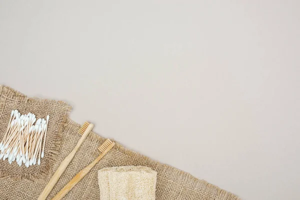 Bamboo toothbrushes, organic loofah, cotton swabs and brown sackcloth on grey background — Stock Photo