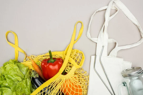 Bolsa de hilo amarillo con verduras frescas maduras, paja de metal, tarro de vidrio y bolsa de algodón sobre fondo gris - foto de stock