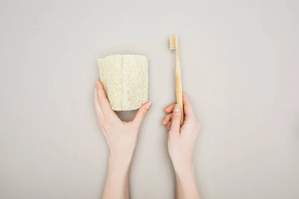 Vue recadrée d'une femme tenant une éponge organique et une brosse à dents en bambou sur fond gris — Photo de stock