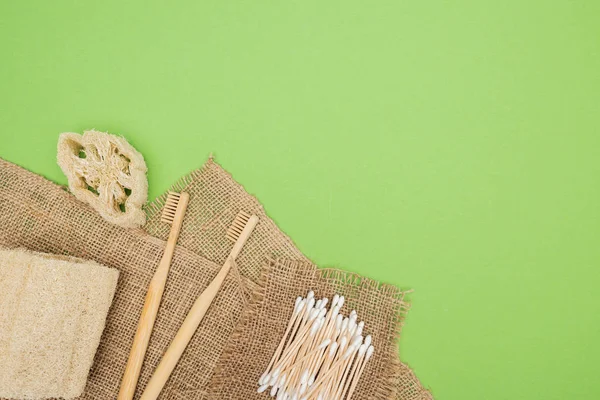 Bamboo toothbrushes, organic loofah, cotton swabs and brown sackcloth on light green background — Stock Photo