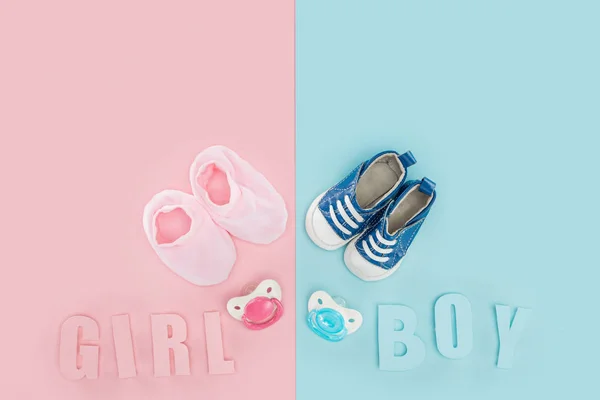 Draufsicht auf Schnuller, Booties, Turnschuhe und Jungen, Mädchen Schriftzug auf rosa und blauem Hintergrund — Stockfoto