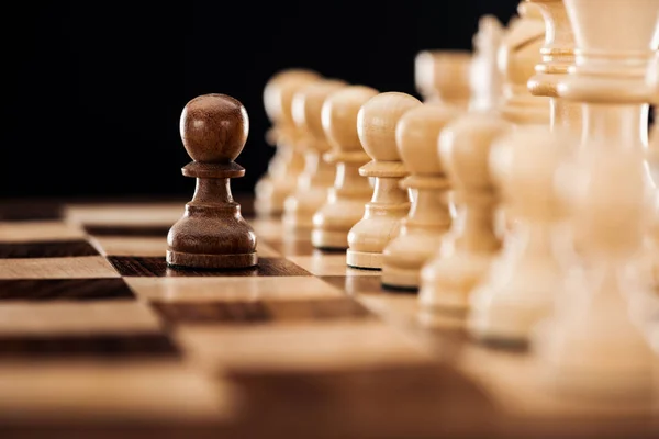 Selective focus of wooden chessboard with beige chess figures and brown pawn in front isolated on black — Stock Photo