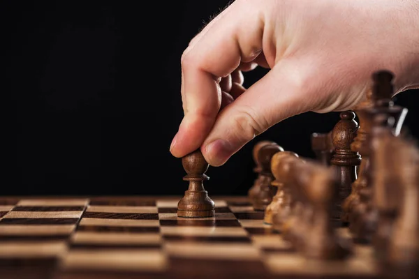 Visão cortada do homem fazendo movimento com peão no tabuleiro de xadrez de madeira isolado no preto — Fotografia de Stock