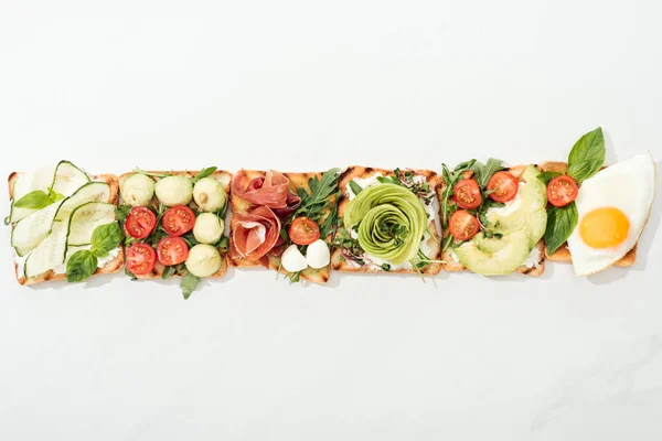 Top view of toasts with cut vegetables, fried egg and prosciutto on white surface — Stock Photo