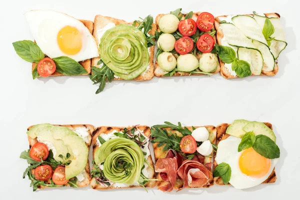 Top view of toasts with cut vegetables, fried eggs and prosciutto on white surface — Stock Photo