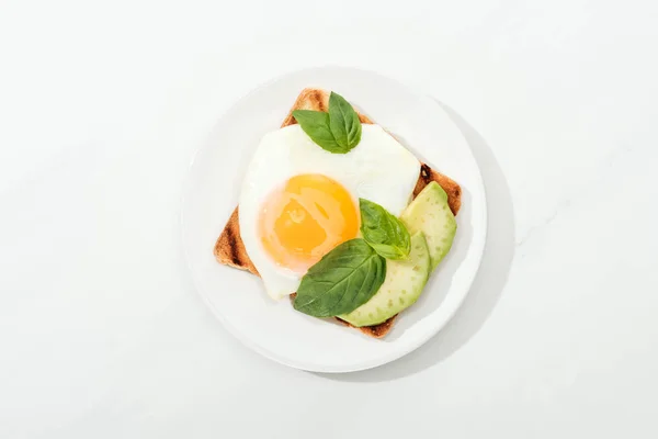 Вид зверху на тост зі смаженим яйцем і нарізаним авокадо на тарілці на білій поверхні — стокове фото
