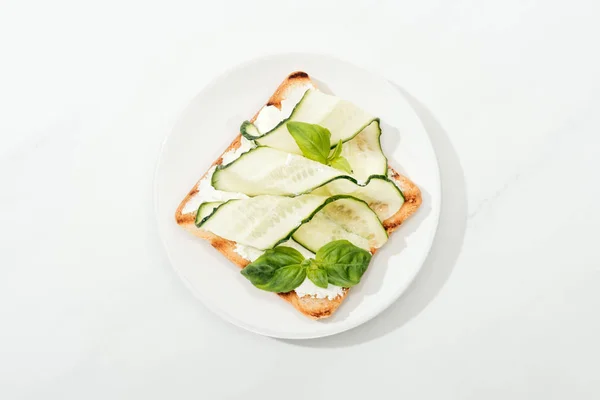Вид зверху на тост з нарізаним огірком і базиліком на тарілці на білій поверхні — стокове фото