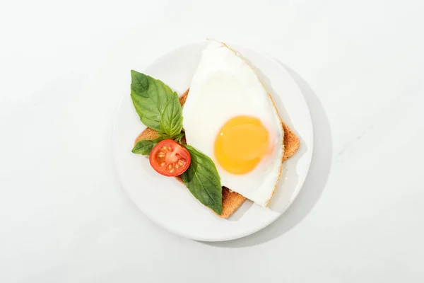 Top view of toast with fried egg, basil and cut tomato cherry on plate on white surface — Stock Photo