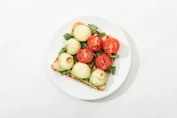 Вид зверху на тост з вишневими помідорами та руколою на тарілці на білій поверхні — стокове фото