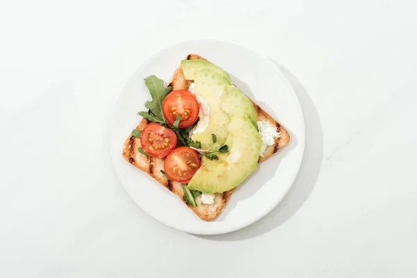 Vista dall'alto di pane tostato con pomodorini ciliegia e avocado su piatto su superficie bianca — Foto stock