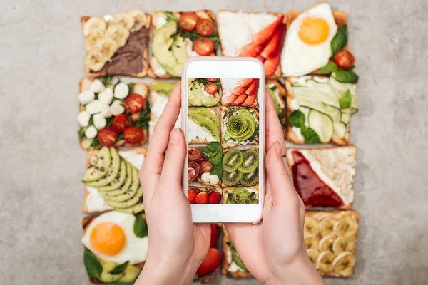 Partial view of woman holding smartphone and taking photo of toasts with fruits and vegetables — Stock Photo