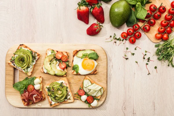 Top view of toasts with vegetables on chopping board and fresh ingredients on wooden table — Stock Photo