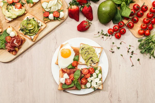 Top view of ingredients and toasts with vegetables, fried egg and prosciutto on wooden table — Stock Photo