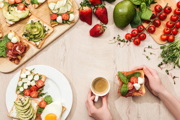 Draufsicht der Frau mit einer Tasse Kaffee am Tisch mit Zutaten, Grün und Toasts mit Gemüse und Prosciutto — Stockfoto