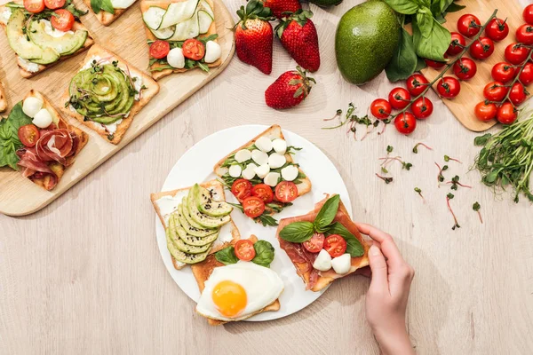 Vista superior de la mujer con deliciosas tostadas de verduras e ingredientes en la mesa de madera - foto de stock