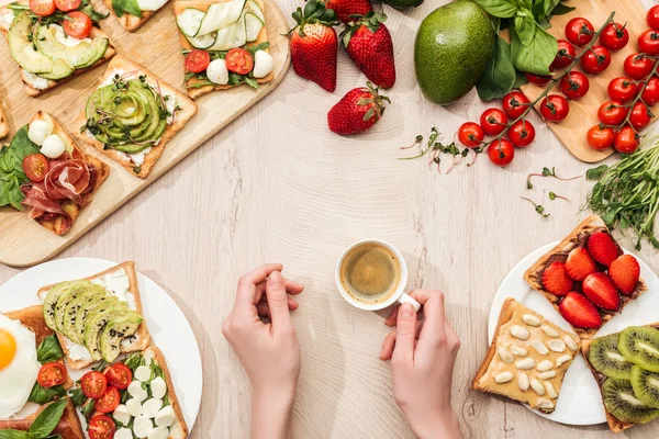 Top view of woman holding cup of coffee at table with fresh ingredients, greenery and toasts with vegetables and prosciutto — Stock Photo