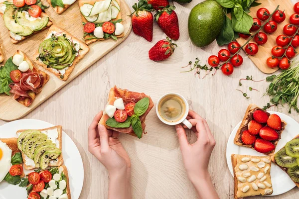 Top view of woman holding cup of coffee at table with ingredients, greenery and toasts with vegetables and prosciutto — Stock Photo