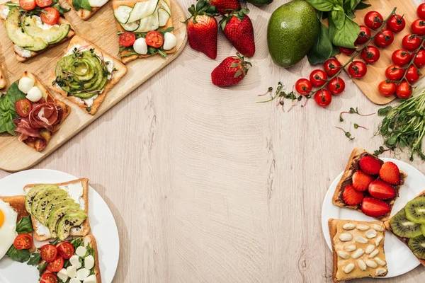 Vista superior de torradas com legumes, frutas e prosciutto com vegetação e ingredientes em mesa de madeira — Fotografia de Stock