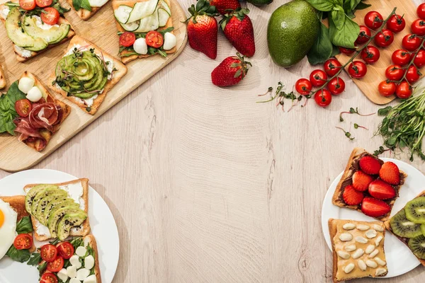 Top view of toasts with vegetables, fruits and prosciutto with greenery and ingredients on wooden table — Stock Photo