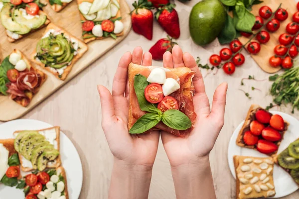 Selective focus of woman holding toast with cherry tomatoes and prosciutto over wooden table with greenery and ingredients — Stock Photo