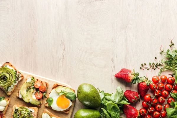 Top view of ingredients on wooden table and toasts with vegetables on chopping board — Stock Photo
