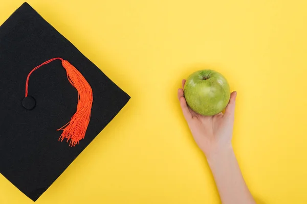 Cropped view of woman with academic cap holding green apple on yellow surface — Stock Photo