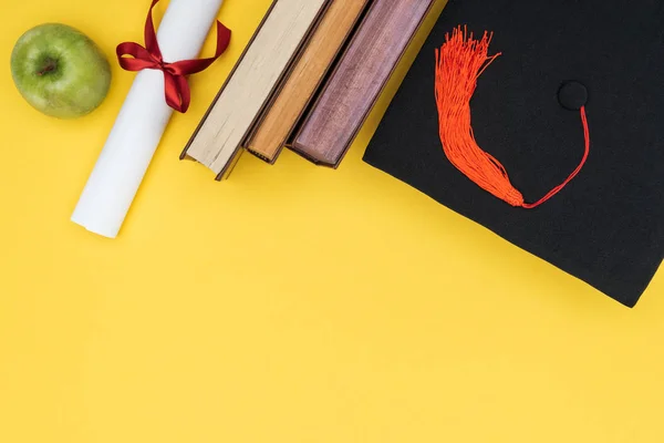 Вид зверху на книжки, яблуко, академічна шапка та диплом на жовтій поверхні — стокове фото