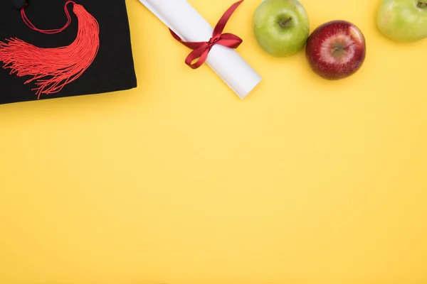 Vista superior de gorro acadêmico, diploma e maçãs na superfície amarela — Fotografia de Stock