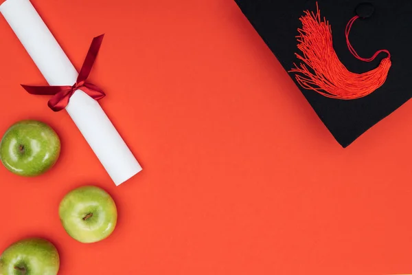 Vista superior de la tapa académica, diploma y manzanas en la superficie roja - foto de stock