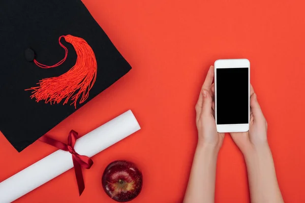 Vista parcial de la chica con gorra académica y diploma usando smartphone con pantalla en blanco en la superficie roja - foto de stock