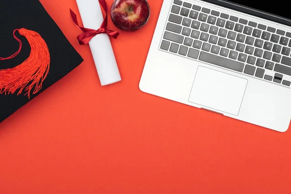 Vue du dessus de l'ordinateur portable, diplôme, casquette académique et pomme sur la surface rouge — Photo de stock