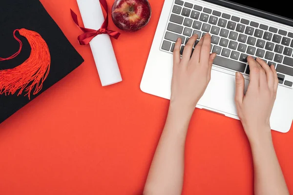 Cropped view of girl with academic cap, diploma and apple using laptop on red surface — Stock Photo