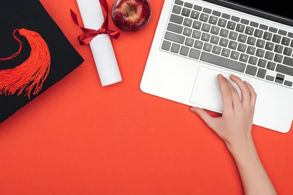 Cropped view of girl with academic cap, diploma and apple using laptop on red surface — Stock Photo