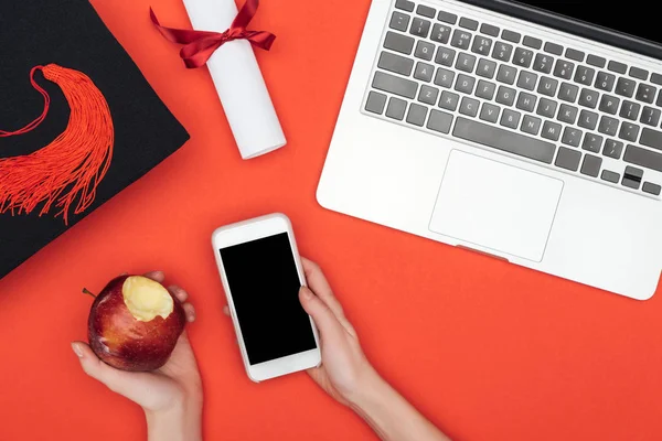 Cropped view of student holding apple and smartphone with blank screen on red surface — Stock Photo