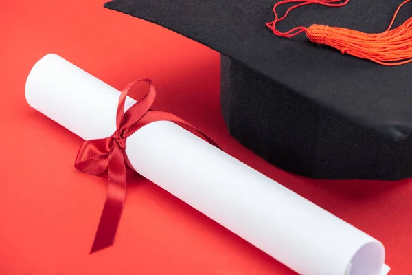 Tampa acadêmica com borla e diploma com fita na superfície vermelha — Fotografia de Stock