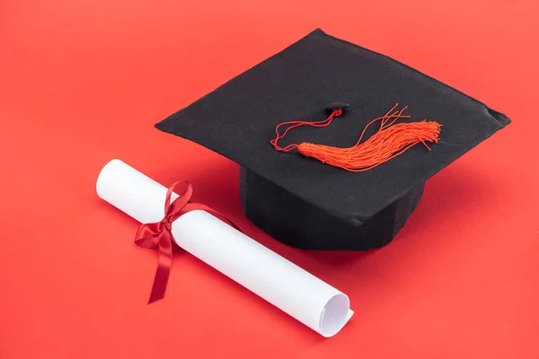 Tampa acadêmica com borla e diploma com fita na superfície vermelha — Fotografia de Stock