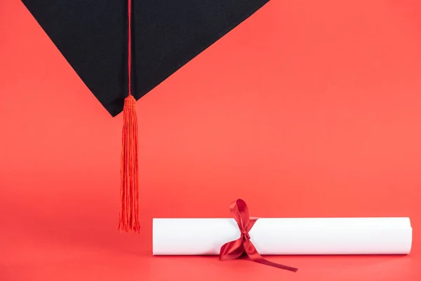 Casquette académique avec pompon et diplôme avec ruban sur surface rouge — Photo de stock