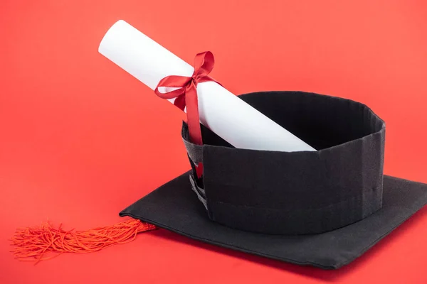 Casquette académique avec pompon et diplôme avec ruban sur surface rouge — Photo de stock