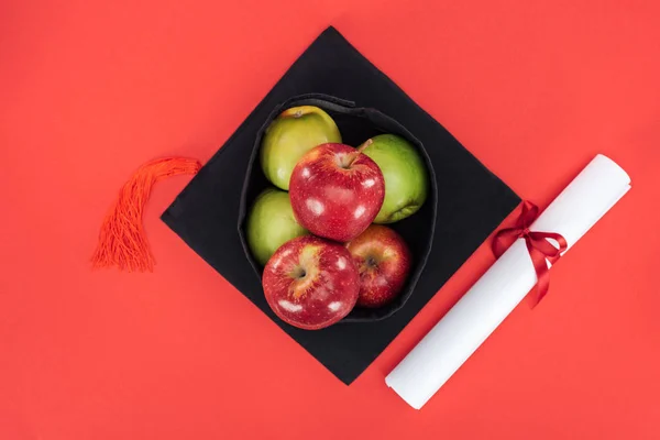 Vista superior da tampa acadêmica com maçãs e diploma na superfície vermelha — Fotografia de Stock