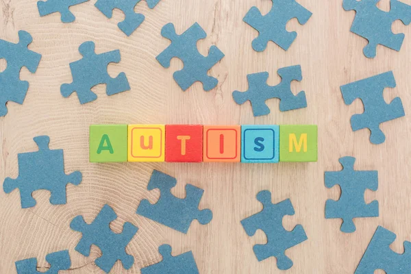 Vista superior de letras autismo feito de cubos multicoloridos entre peças de quebra-cabeça azuis na mesa de madeira — Fotografia de Stock